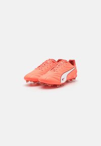 Des crampons de football corail vif avec une tige synthétique texturée, arborant un design à rayures blanches et des pointes moulées pour une traction sur les surfaces en herbe.