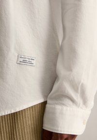 Biała bawełniana koszula z długim rękawem i mankietem zapinanym na guziki, z naszywką z tekstem. Koszula ma gładką teksturę i klasyczny krój.