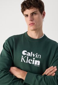 Felpa verde con colletto rotondo, caratterizzata da testo bianco "Calvin Klein" ben visibile sul davanti. Tessuto morbido, vestibilità casual.
