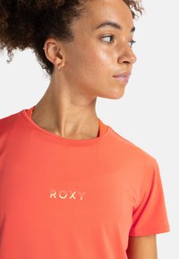 Camiseta atlética color coral de manga corta, con cuello redondo y detalle del logo "ROXY" en dorado. Textura suave con un diseño ajustado.