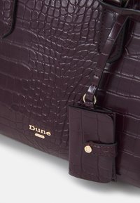 Fioletowa torebka z wytłoczeniem w kształcie krokodyla, o gładkiej teksturze, z logo w kolorze złotym i małą odpinaną kieszonką przymocowaną paskiem.