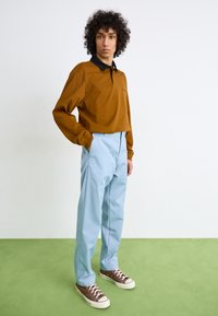 Brun lange erme polo med sort krave, parret med lyseblå bukser og brune sneaks, sat mod en neutral baggrund.
