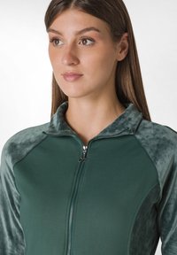 Giacca sportiva verde con zip frontale, caratterizzata da un corpo testurizzato e maniche lisce e vellutate. Collo e dettagli in una tonalità coordinata.