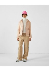 Giacca a vento rosa chiaro con cuciture orizzontali, dolcevita bianco, pantaloni beige a vestibilità ampia con strisce laterali a contrasto e sneakers argentate.