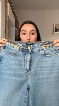 Γυναίκα κρατάει ανοιχτό γαλάζιο τζιν με χρυσή αλυσίδα, δείχνοντας τη μέση και τις μπροστινές τσέπες, σε εσωτερικό χώρο με ουδέτερο φόντο.