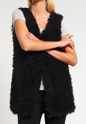 Personne portant un gilet noir sans manches et duveteux sur un t-shirt gris à manches courtes, les bras croisés au niveau de la poitrine.