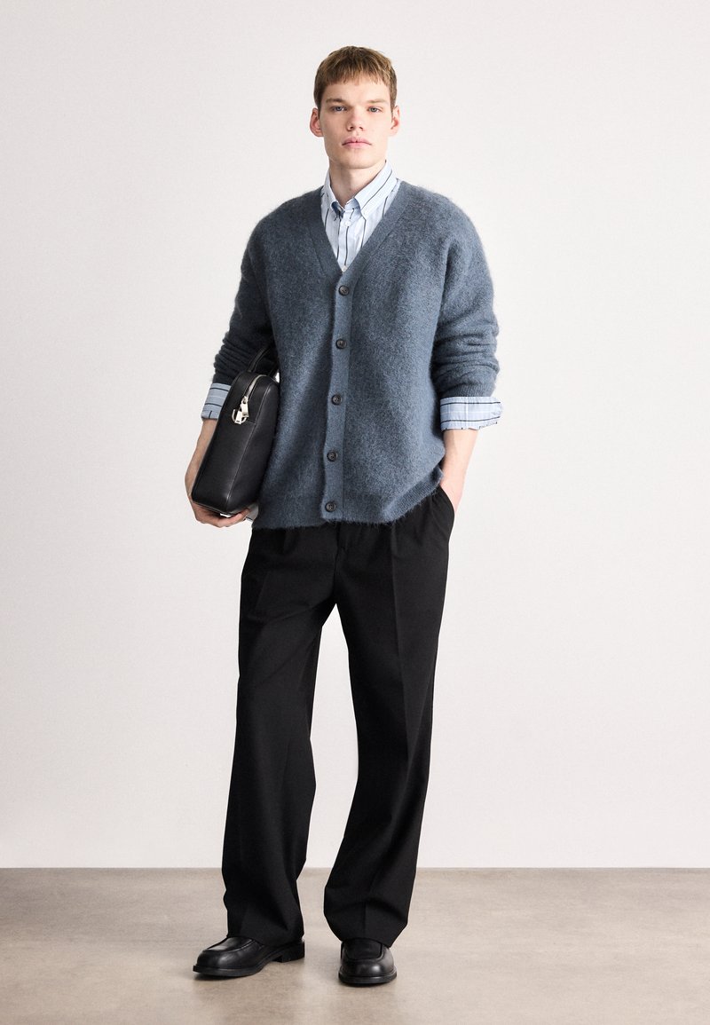 Light blue mohair cardigan with a v-neck and five buttons, paired with a striped shirt and dark trousers. Black structured bag in hand.