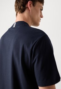 Mørkeblå bomulls t-skjorte med korte ermer, med en ribbestrikket krage og to hvite striper ved bakhalsen. Glatt tekstur, uformell design.