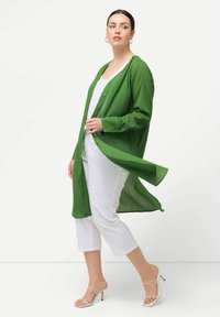 Green lightweight button-up shirt with a flowing design, worn over a white top and white cropped pants. The model wears clear strappy heels.