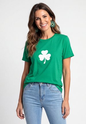 Kobieta uśmiechnięta, ubrana w zielony t-shirt z białym wzorem koniczyny i niebieskie dżinsy, z zielonymi ozdobnymi kolczykami, stojąca na białym tle.