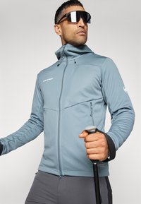 Lyseblå atletisk jakke med en tætsiddende design, lynlåslommer og høj krave, parret med mørke bukser og holder en trekkingstok.