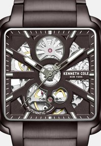 Quadratische automatische Uhr mit einem dunkelbraun strukturierten Zifferblatt, transparenten Abschnitten, silbernen Akzenten und auffälligen Zahnrädern. Das Armband hat ein gebürstetes Metall-Finish.