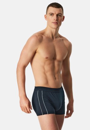 Man zonder shirt met kort haar, gekleed in donkerblauwe boxershorts met witte zijstrepen, staand tegen een effen lichte achtergrond.