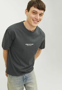 Camiseta de algodón gris oscuro con mangas cortas y cuello redondo, con el texto blanco "Originals Studio" en el frontal. Jeans de color azul claro.