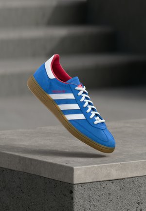 Μπλε αθλητικό παπούτσι Adidas με λευκές ρίγες, λαστιχένια σόλα και κόκκινη εσωτερική επένδυση, τοποθετημένο στον αέρα πάνω από σκυρόδεμα.