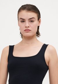 Juodas, raukšlėtas tank top'as su kvadratine iškirpte ir plačiomis petnešomis, pasižymintis prigludusia siluetu ir švelnia tekstūra.