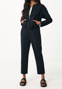 Svart jumpsuit med dragkedja framtill, elastisk midja, långa ärmar och tapered ben i ankellängd, tillsammans med svarta flätade sandaler.