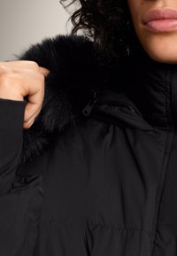 Čierna nadýchaná bunda s umelým kožušinovým golierom, so zatváraním na zips, jemnou textúrou a prešívaným dizajnom zdôrazňujúcim teplo a funkčnosť.