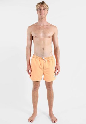 Homme blond debout pieds nus, face à l’avant, portant un short de bain couleur pêche avec un cordon blanc et une petite broderie latérale.