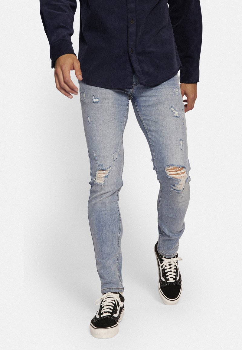 Jeans de mezclilla azul claro con un ajuste slim, que presentan zonas desgastadas y detalles de desgarro. Combinados con zapatillas de cordones en blanco y negro.