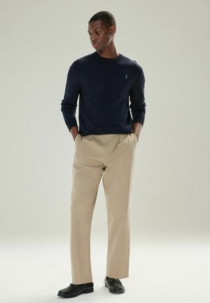 Homme portant un pull bleu marine, un pantalon beige et des mocassins noirs, se tenant les mains dans les poches contre un fond uni.