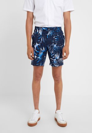 Homme portant un short bleu et noir à motif feuilles tropicales, une chemise blanche à manches courtes et des baskets blanches, se tenant devant un fond uni.