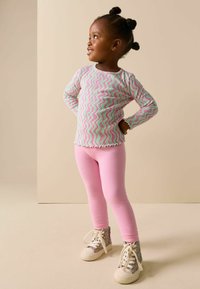 Bluză cu mâneci lungi, cu model tip dungi și valuri multicolore; colanți roz; pantofi sport înalți și masivi, cu model, șireturi și talpă albă.