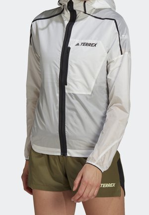 Henkilö, jolla on valkoinen Adidas Terrex -takki mustalla vetoketjulla ja vihreät Terrex-shortsit, seisoo yksiväristä taustaa vasten.