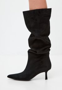 Black knee-high boots made of soft suede, featuring a slouchy design, pointed toe, and a slim stiletto heel. Smooth texture throughout.