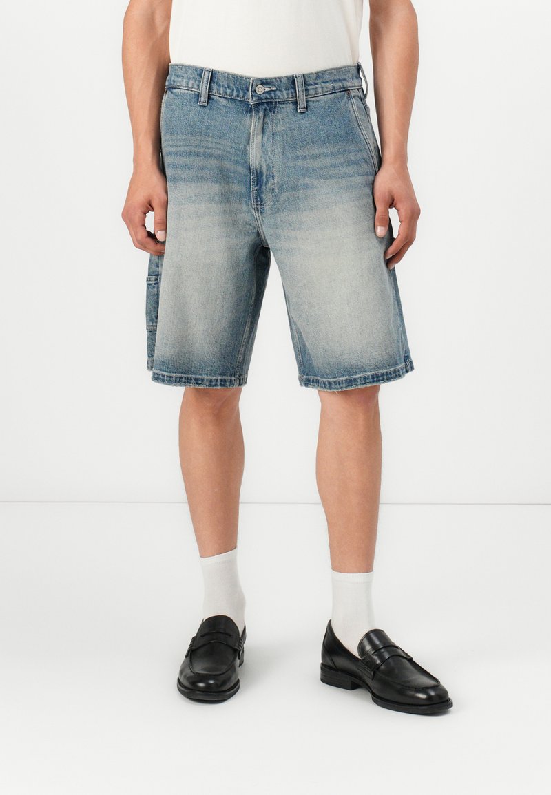 Shorts de mezclilla con un lavado azul desgastado, hasta la rodilla, con bolsillos frontales y un dobladillo ligeramente deshilachado. Combinados con mocasines negros y calcetines blancos.