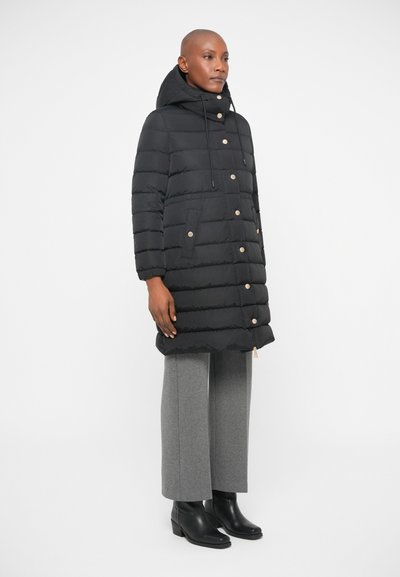 Black puffer coat with a hood, featuring horizontal stitching, two front pockets, gold accents, and a zip closure. Worn with gray trousers and boots.