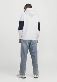 Hvid hættebluse med marineblå accenter, afslappet pasform, ribbede manchetter og bund; parret med lyseblå jeans og grå træningssko. Bagud fra vinkel.