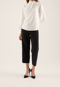 Femme portant un chemisier blanc à manches longues, un pantalon noir coupé à la cheville et des escarpins noirs pointus, debout devant un fond beige.