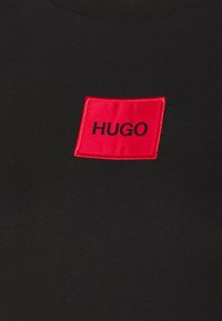 T-shirt noir avec un patch rectangulaire rouge portant le texte noir "HUGO" au centre. Texture de tissu lisse, design standard à col rond.