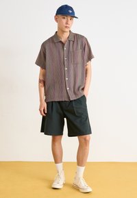 Short-sleeve, multi-colored striped shirt with a chest pocket. Paired with dark shorts and white socks, and a blue cap.