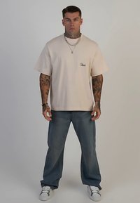 Beige bomulls-t-shirt med korta ärmar och en liten logotyp på bröstet, kombinerad med blå, löst sittande jeans och vita sneakers.