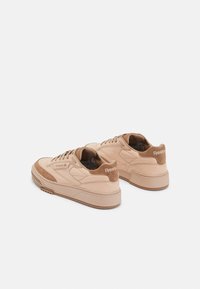 Beige suede sneakers with rounded shape, featuring a textured sole, lace-up design, and embossed logo on the back. Similar tones throughout.