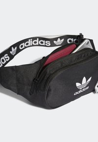Musta kangaskotelo vyölaukku vetoketjulla, jossa on viininpunainen vuori ja valkoinen Adidas-logo edessä ja hihnassa.