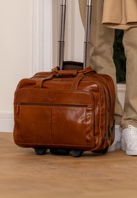 Brown leather rolling suitcase on wooden floor beside person wearing beige pants and white sneakers.