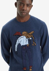 Marineblå strikkegenser med cowboygrafikk, detaljert med brun hatt, hvit skjorte og blå jeans. Rund hals, myk tekstur.