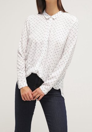 Kobieta ubrana w białą koszulę z długim rękawem z małymi kwiatowymi wzorami i ciemnoniebieskie dżinsy, ręce złożone z przodu.