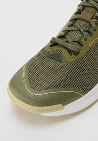 Nike bėgimo bateliai su tekstūruota alyvuogių žalia viršūne, turintys sustiprintą pirštinę ir lengvą baltą pado su dėmėtu apdaila.
