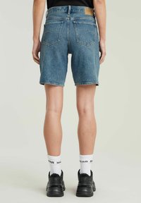 Denim shorts in lichte blauwe wassing met een rechte snit, voorzien van achterzakken en een leren merkpatch. Gecombineerd met zwarte sneakers en witte sokken.
