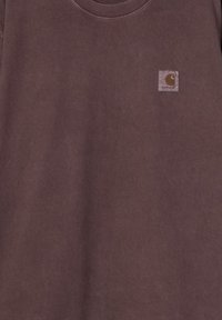 Camiseta de algodón marrón con cuello redondo, que presenta un pequeño parche cuadrado con el logo en la esquina superior izquierda. Textura suave, diseño casual.