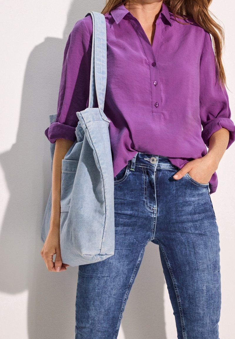 Femme portant une blouse violette à manches longues et un jean bleu avec un sac en tissu bleu clair sur l’épaule.
