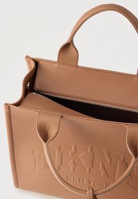 Sac fourre-tout DKNY en cuir texturé marron clair avec double poignées, compartiment supérieur à fermeture éclair ouverte et logo en relief sur le devant.
