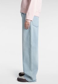 Pantaloni larghi in denim azzurro chiaro con vita alta, caratterizzati da una singola tasca posteriore e una texture morbida. Indossati con scarpe a fantasia.