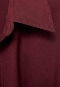 Gros plan sur un col de chemise rouge foncé montrant la texture du tissu et les coutures détaillées sur un matériau en coton lisse.