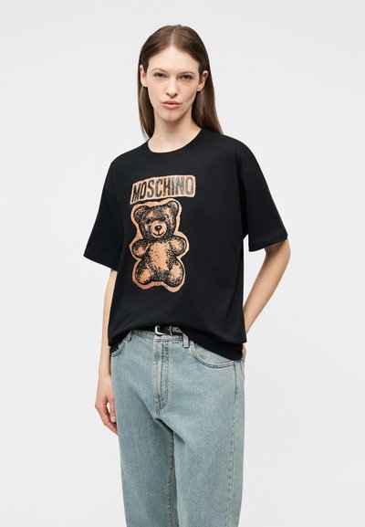 Γυναίκα που φοράει μαύρη μπλούζα Moschino με μεγάλο γραφικό αρκουδάκι και κείμενο, συνδυασμένη με ανοιχτόχρωμο τζιν και μαύρη ζώνη.