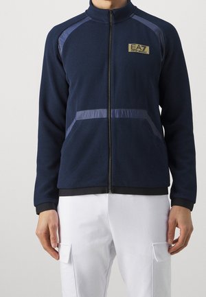 Mand iført navyblå lynlåstrøje med tekstureret stof, reflekterende striber og EA7 Emporio Armani-logo, kombineret med hvide cargobukser.